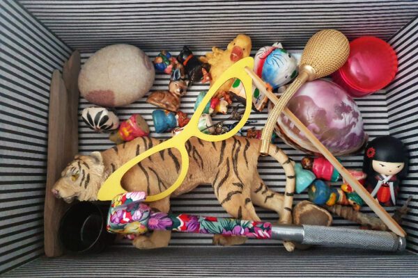 A box containing objects, including a plastic toy tiger, a pair of yellow toy glasses, a hammer with a floral pattern, ceramic toy figures, a shell with African wildlife embossed.