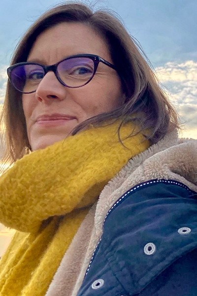 Vicki Lamb headshot image. Vicki wears glasses and has a blue coat with a white trim and a yellow scarf. A blue, cloudy sky can be seen in the background